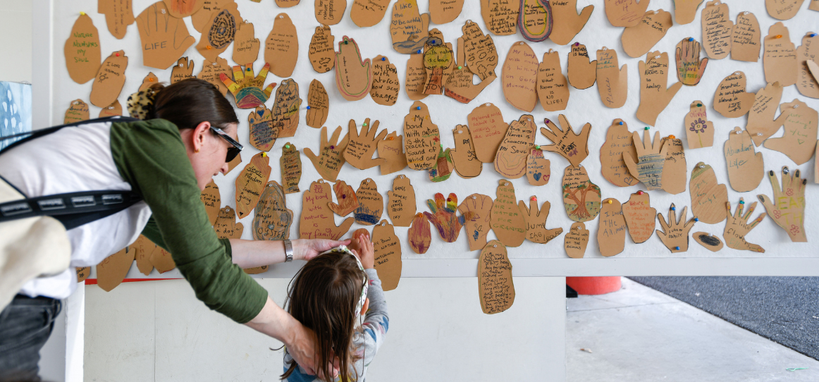 A young girl and mother add paper hands to a community art board