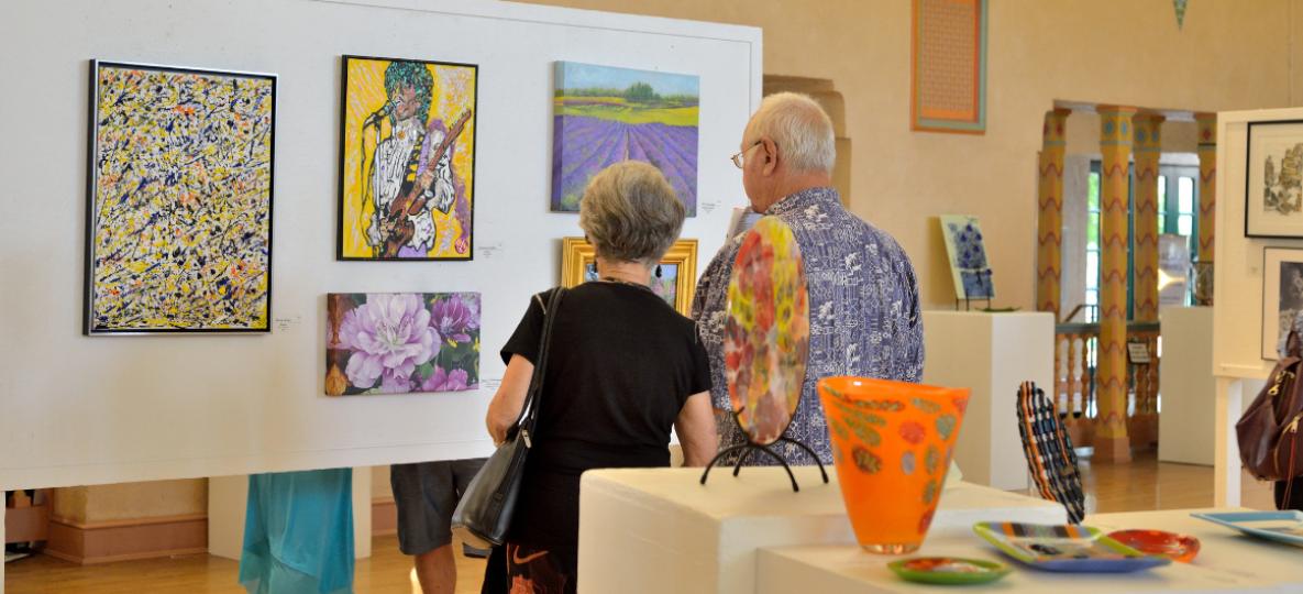 Two people - viewers of the Labor Day Art Show, a man and a woman, both with grey hair - look at the art show.