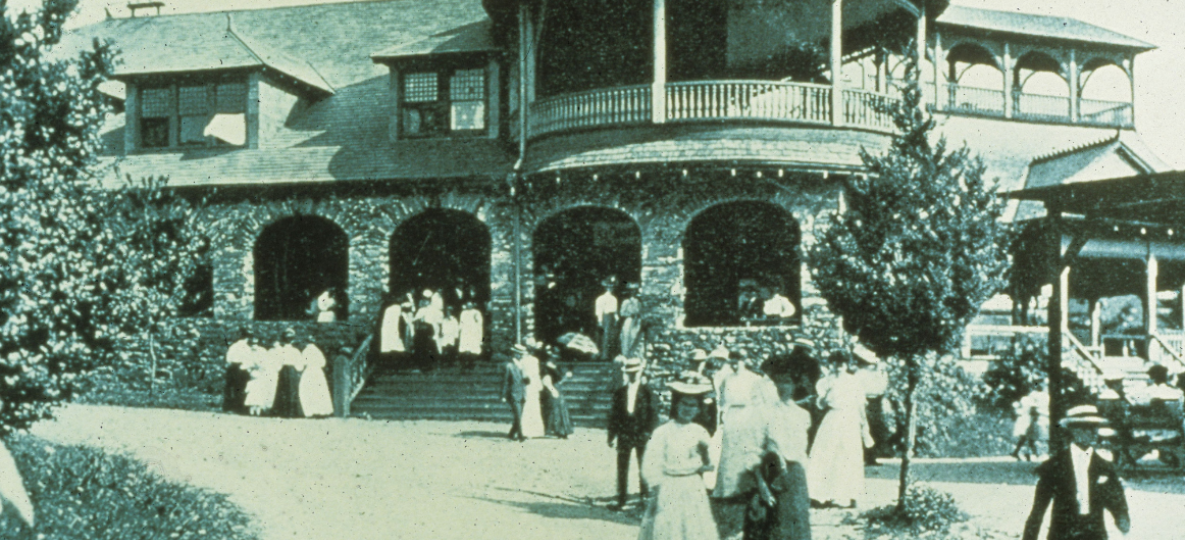 Black and white photo of the Park when it was a Chautauqua in late 1800s with a large stone and wood building and people dressed in late 1800s clothing.